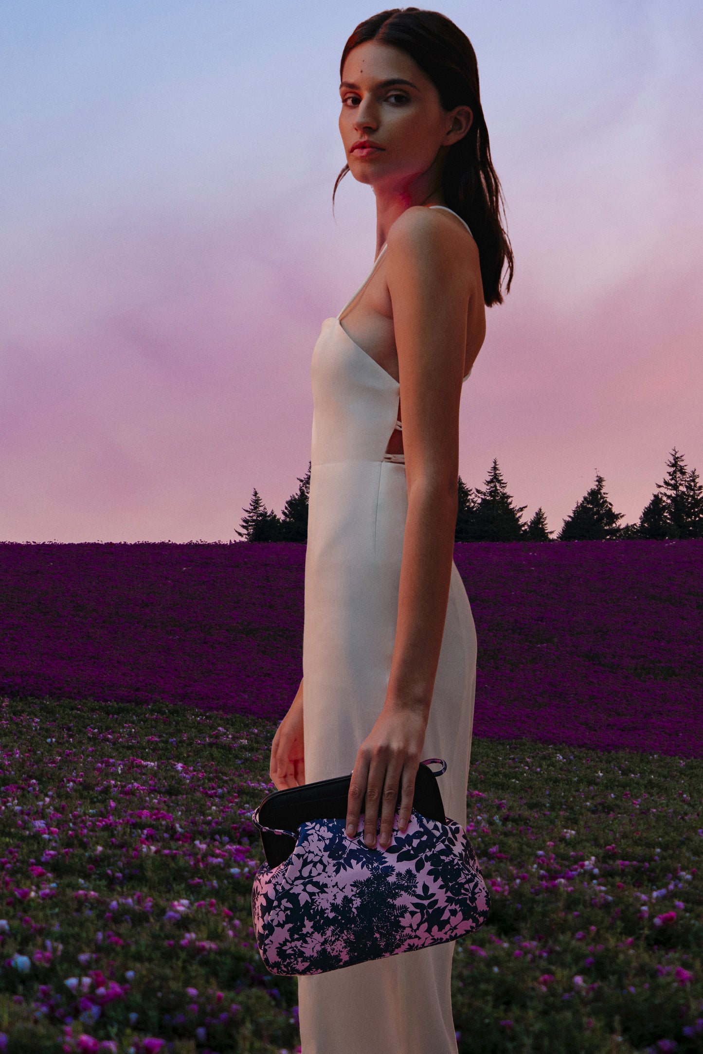 Mujer con vestido blanco en paisaje de lavanda sosteniendo un bolso de mano rosa con hojas azules