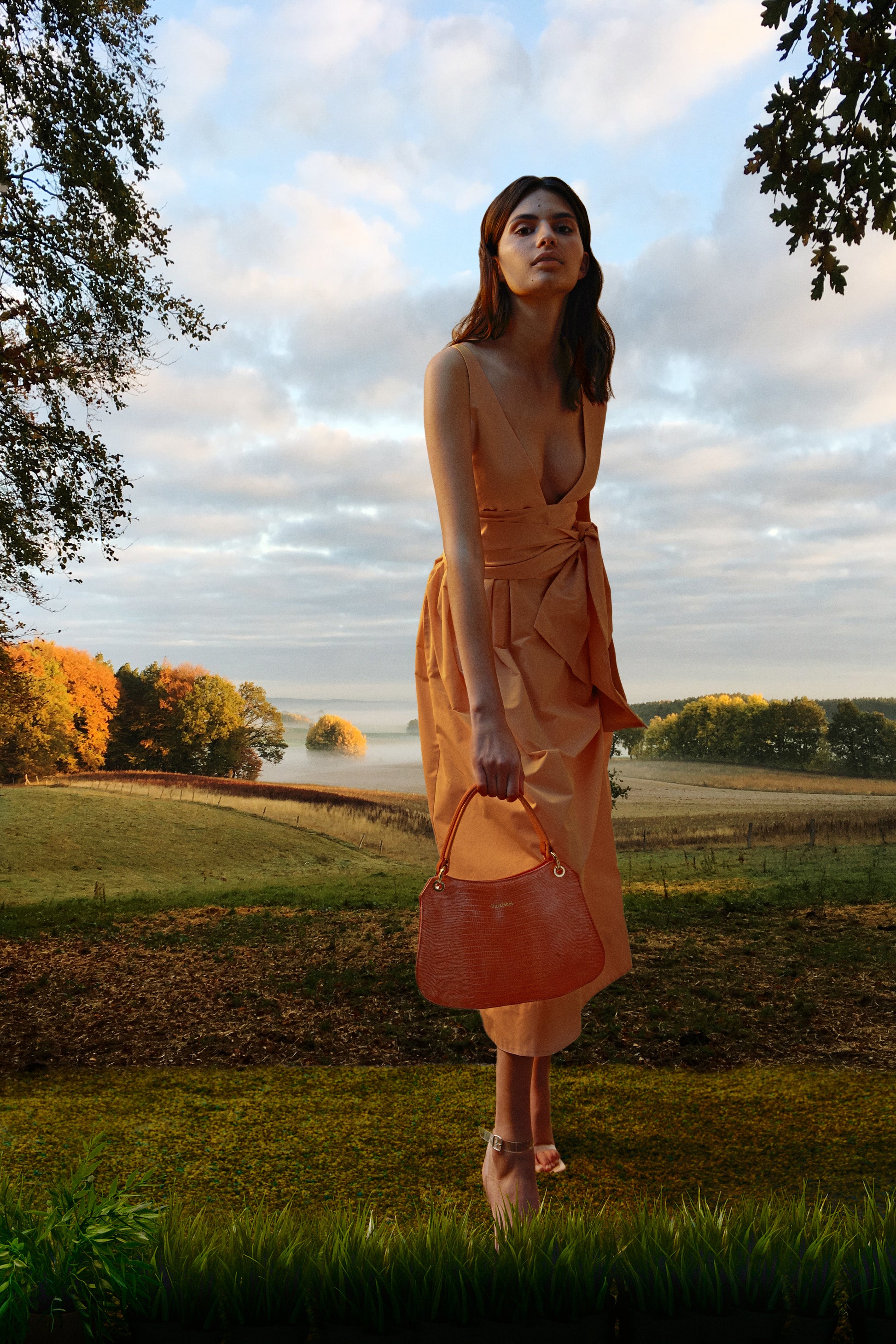 Mujer con paisaje de campo al fondo sosteniendo un bolso naranja metalizado sin bandolera