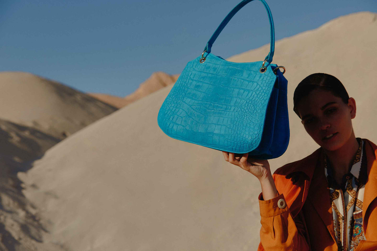 Mujer sosteniendo un bolso de mano azul cocodrilo con unas dunas de fondo