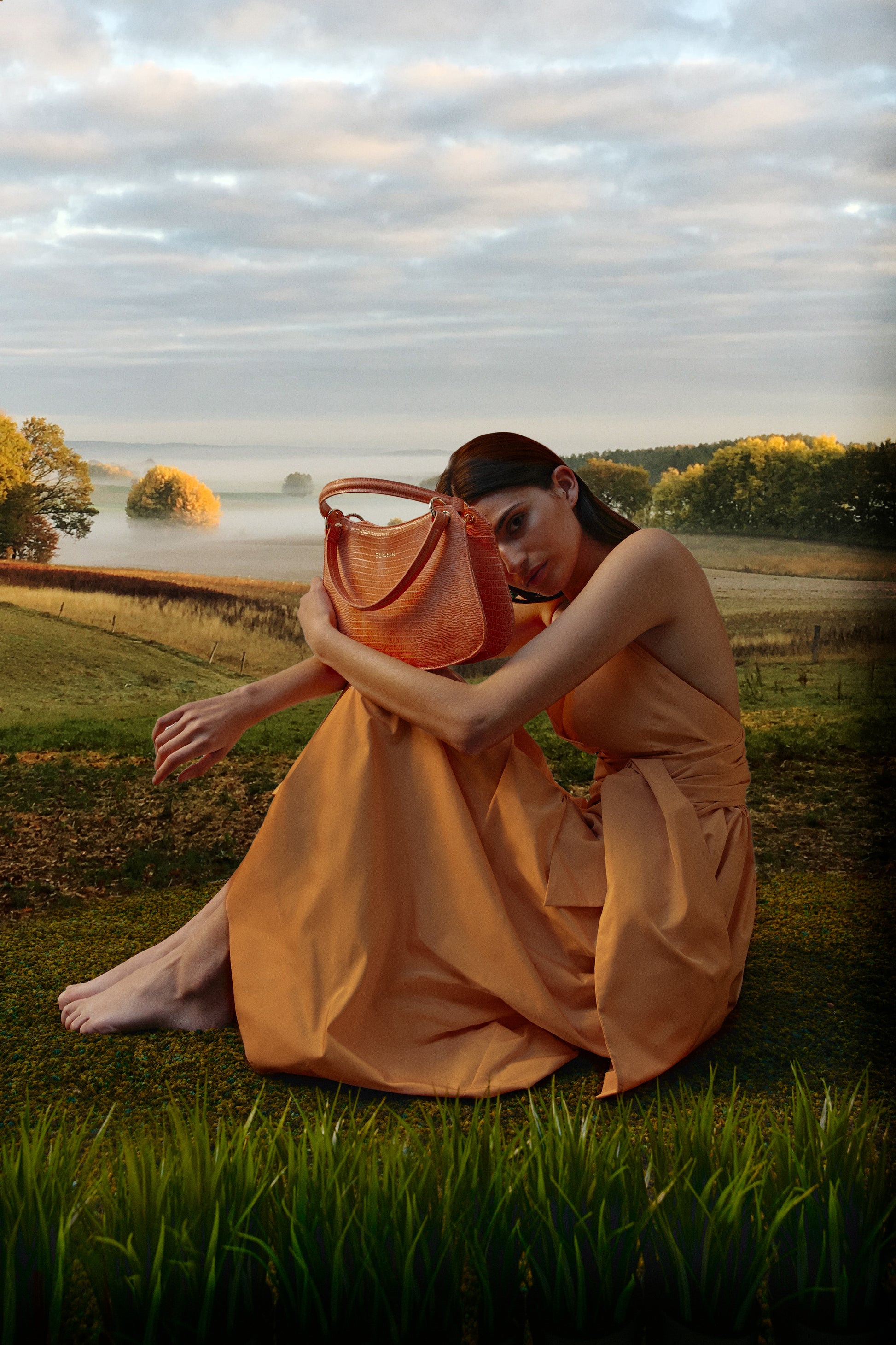 Mujer con vestido naranja sentada sobre un paisaje de naturaleza sosteniendo un bolso de color naranja metalizado efecto lizard